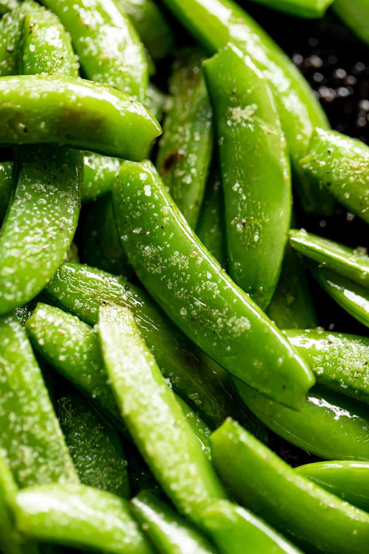 Close up on the green sugar snap peas, sautéed in a black pan, sprinkled with coarse salt.