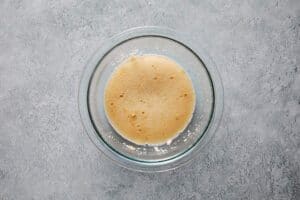 A top-down view of a clear glass bowl filled with a foamy, activated yeast mixture on a gray textured surface.