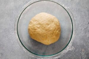 Top-down view of a smooth ball of dough resting in a clear glass bowl covered with plastic wrap on a gray textured surface.