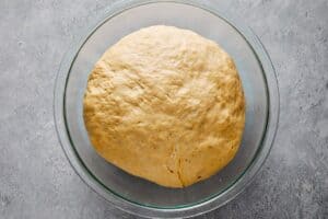 Top-down view of a smooth ball of dough resting in a clear glass bowl covered with plastic wrap on a gray textured surface.