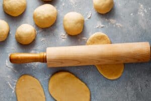 Top-down view of a wooden rolling pin flattening a piece of dough on a flour-dusted gray surface, surrounded by round dough balls.