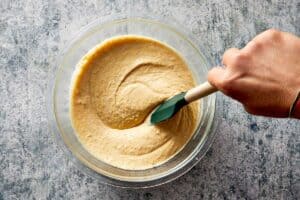 A hand using a green silicone spatula to stir a thick, light brown batter in a clear glass mixing bowl, set on a speckled grey surface.