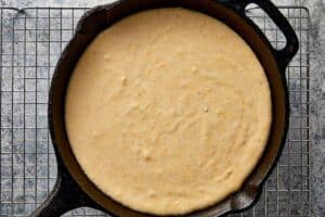 A black cast-iron skillet filled with light brown cornbread batter, resting on a wire cooling rack over a speckled grey surface