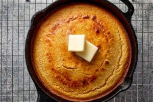 Overhead view of freshly baked, golden-brown cornbread in a black cast-iron skillet, topped with two melting pats of butter and resting on a wire cooling rack.