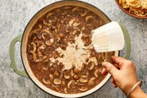 A hand pouring cream from a small pitcher into a skillet of cooked mushrooms and onions.