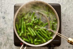 Fresh green beans boiling in a metal pot on a black burner with a wire skimmer spoon
