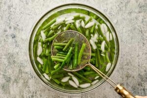 Blanched green beans cooling in a glass bowl filled with ice water and a wire skimmer spoon.