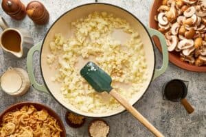 Diced onions sautéing in a large skillet with a spatula surrounded by fresh mushrooms, crispy fried onions and other green bean casserole ingredients.