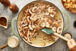 A hand using a green spatula to stir sliced brown mushrooms and diced onions in a large skillet.