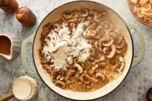 Flour added to a simmering mixture of sautéed mushrooms and onions in a large skillet.