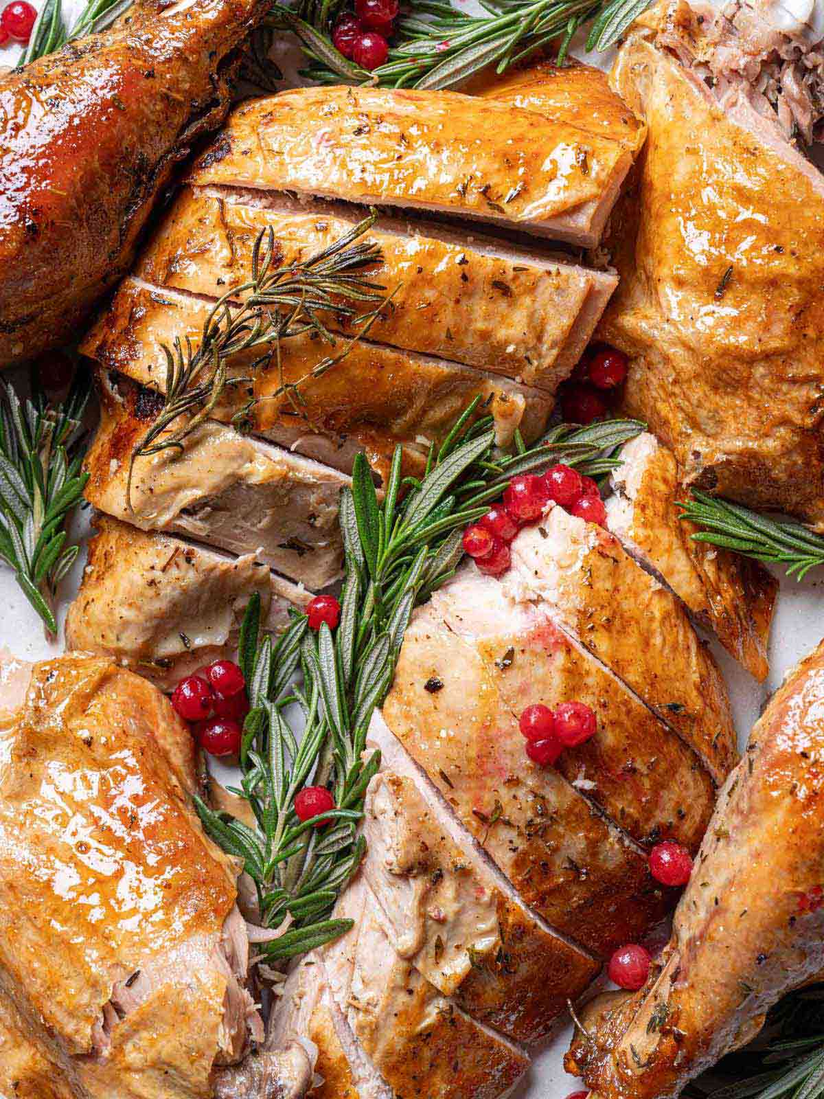 Garlic Herb Butter Roasted Turkey chopped into large slices and pieces, on a serving plate with rosemary sprigs and red cranberries strewn throughout the dish.