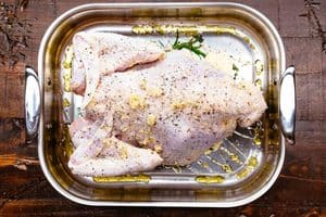 A raw, whole turkey generously seasoned with salt, pepper, and garlic, sitting in a stainless steel roasting pan drizzled with olive oil and fresh herbs on a rustic wooden table.