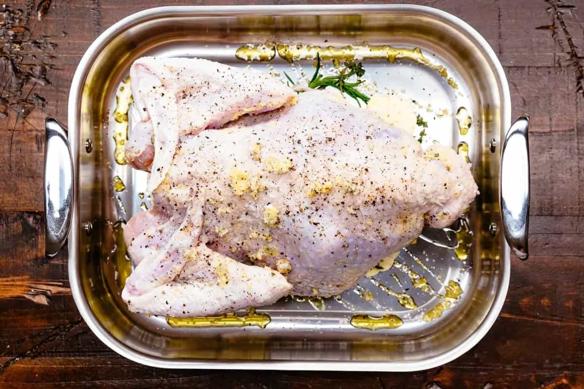 A raw, whole turkey generously seasoned with salt, pepper, and garlic, sitting in a stainless steel roasting pan drizzled with olive oil and fresh herbs on a rustic wooden table.