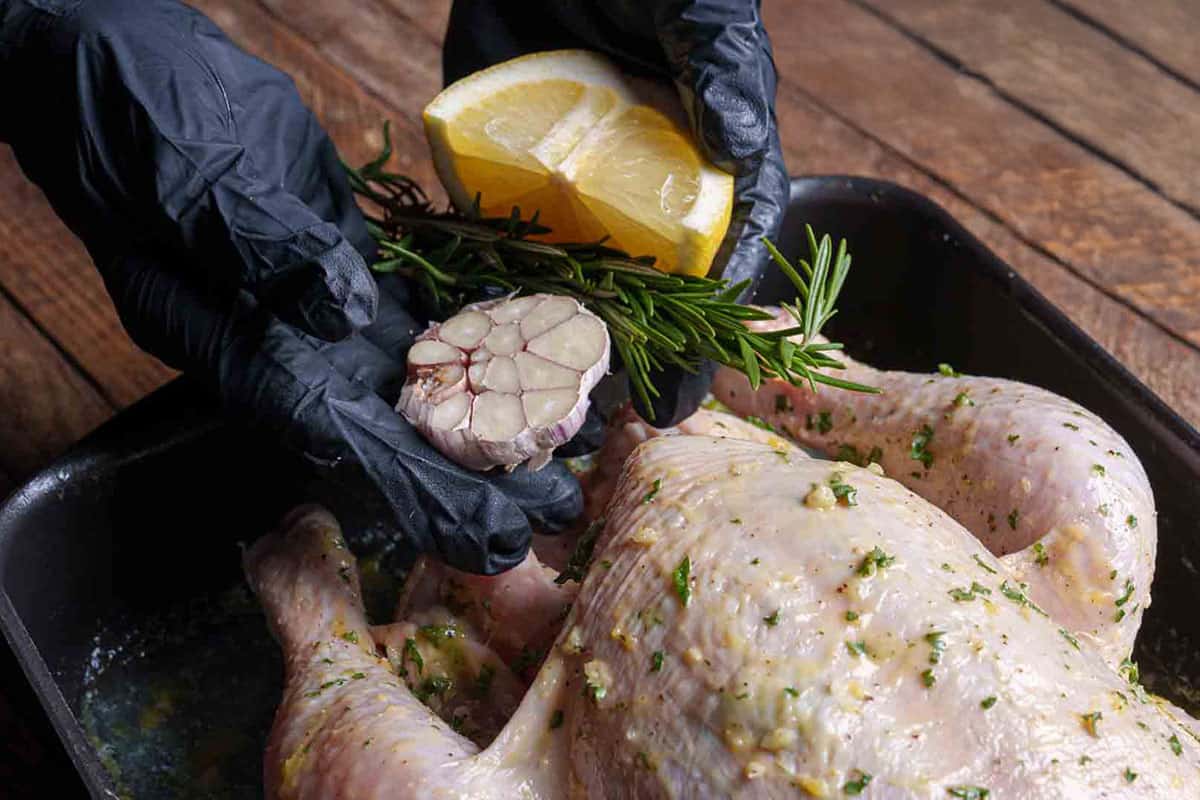 Image showing the chicken being stuffed with a garlic head, rosemary sprigs, and squeezed lemon halves, with legs tied together with kitchen string.