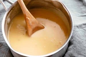 A close-up view of smooth, light-brown turkey gravy in a stainless steel saucepan, with a wooden spoon resting inside it, set against a textured gray cloth background.