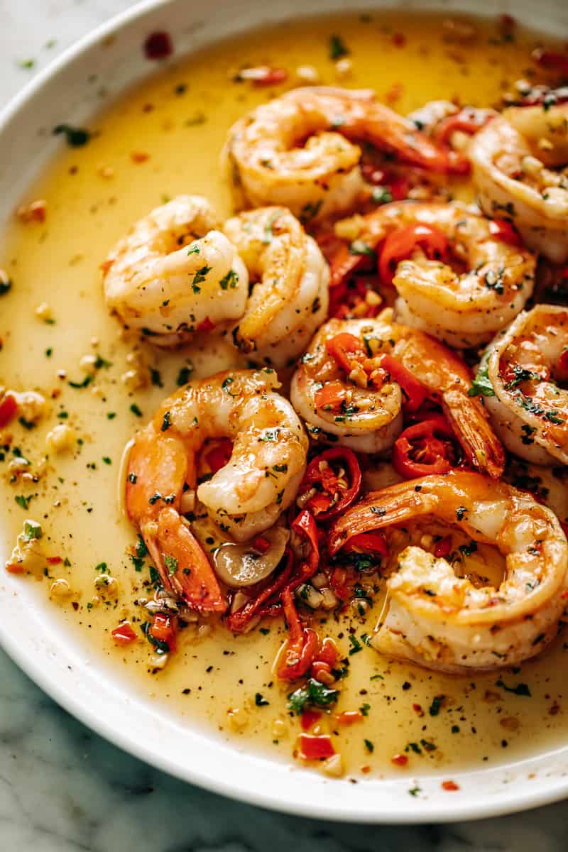 On a white plate, a serving of the chilli garlic butter shrimp in a bed of sauce with pieces of red chilli, green  parsley.