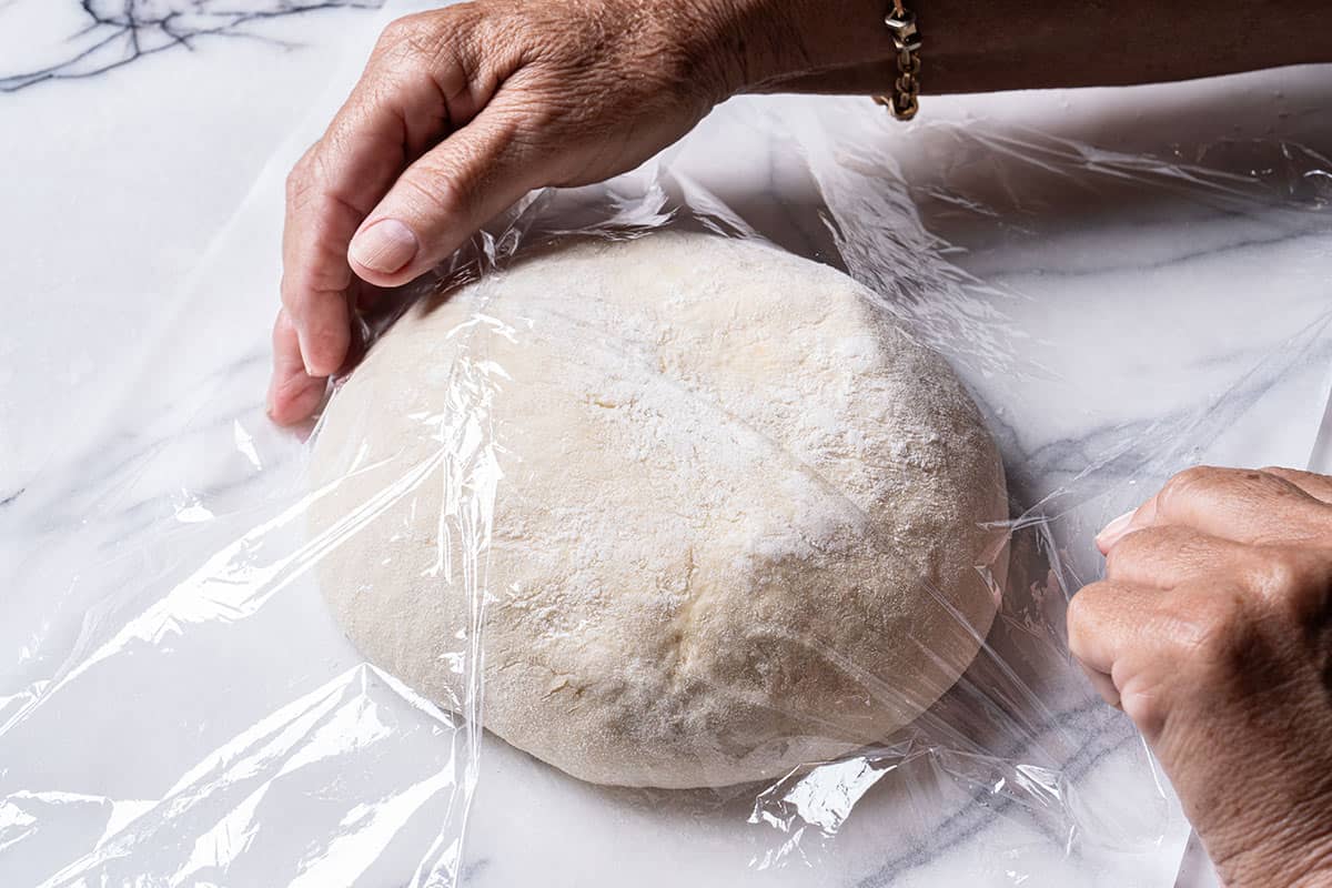 An image of hands covering the dough with parchment paper and then wrapping it in cling film.