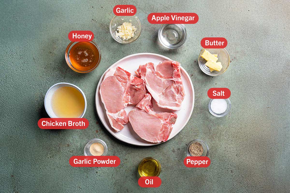 ingredients laid out on a green background in white bowels, honey, garlic, apple vinegar, butter, salt, pepper, oil, garlic powder, chicken broth and pork chops