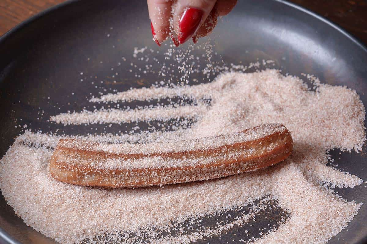 A churro is being removed from the frying pan and dipped into sugar and cinnamon.