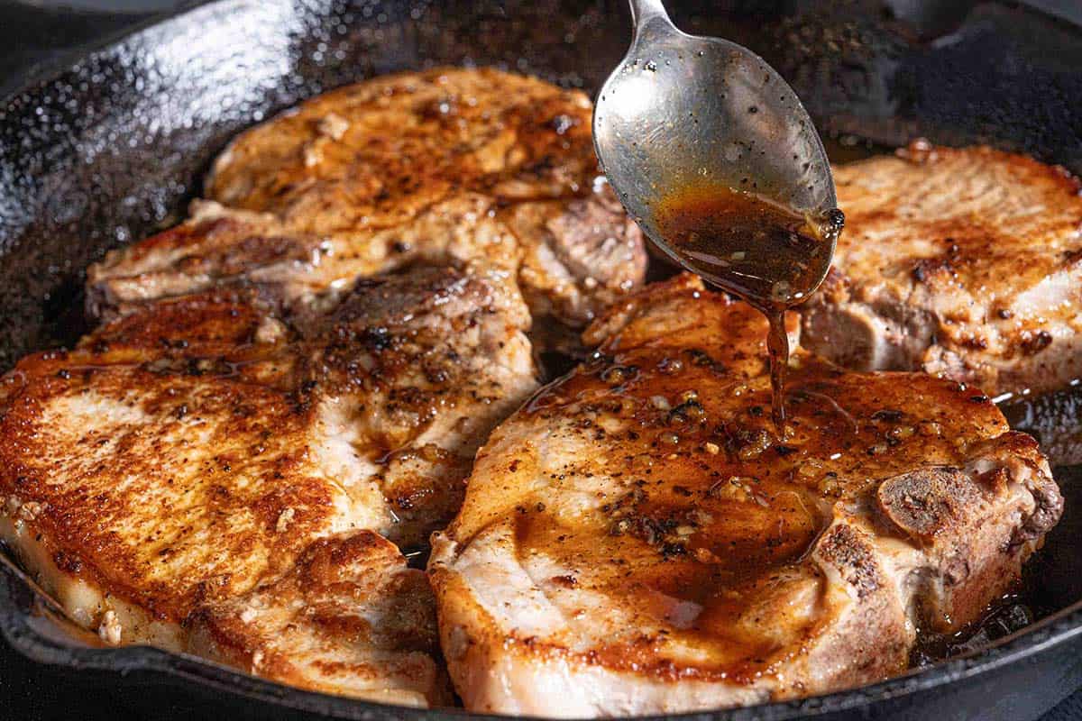 pork chops sizzling in a black pan being doused in honey garlic sauce with a silver spoon