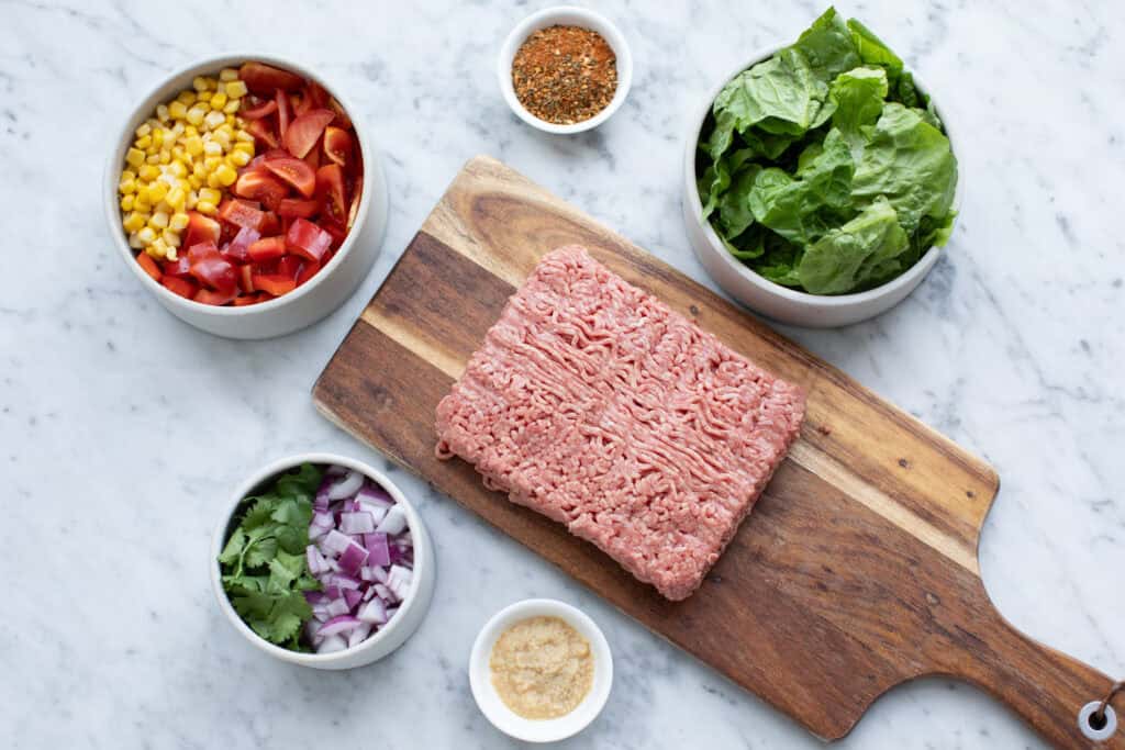 Bird's eye view of raw mince on wooden chopping block, tomatoes, lettuce, corn, spices, onion and coriander in bowls