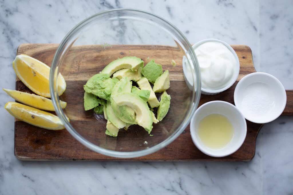 Sliced avocado in glass bowl on wooden board next to lemon, sour cream and other condiments.