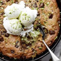 Close-up image of the spoon lifting a portion of pistachio cookie from a skillet, served with vanilla ice cream and topped with chocolate.