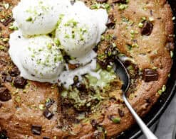 Close-up image of the spoon lifting a portion of pistachio cookie from a skillet, served with vanilla ice cream and topped with chocolate.