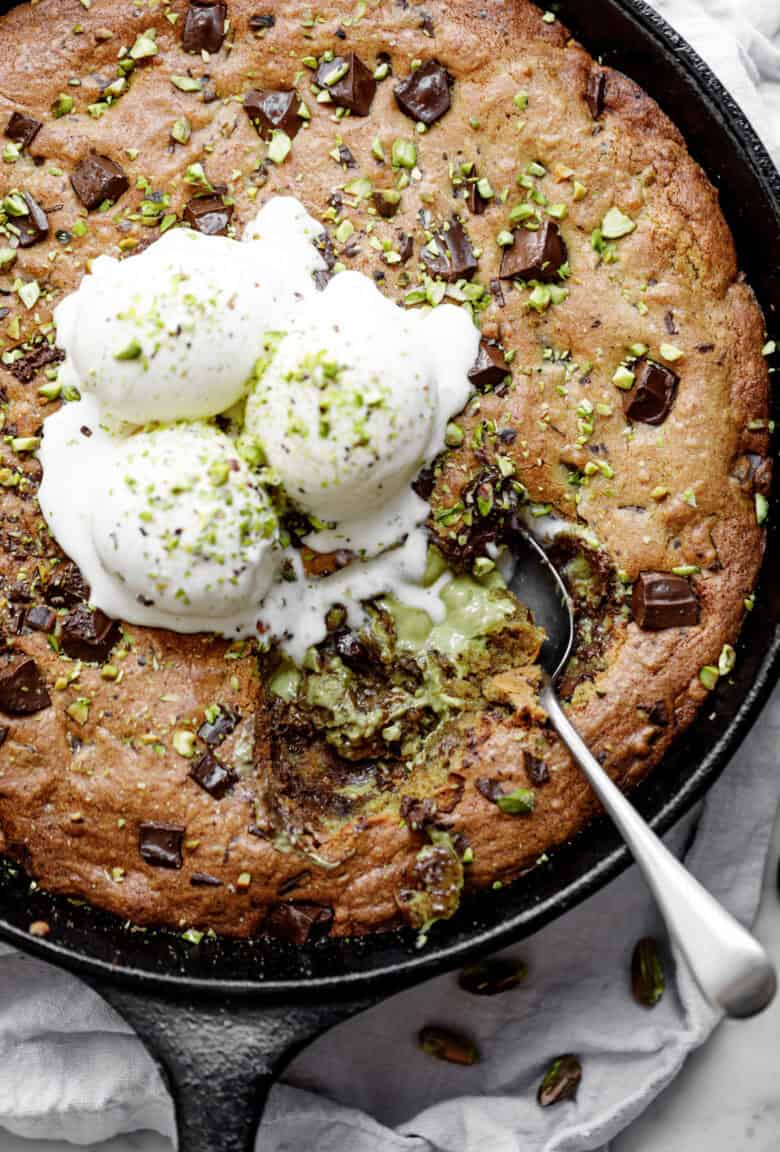 Close-up image of the spoon lifting a portion of pistachio cookie from a skillet, served with vanilla ice cream and topped with chocolate.