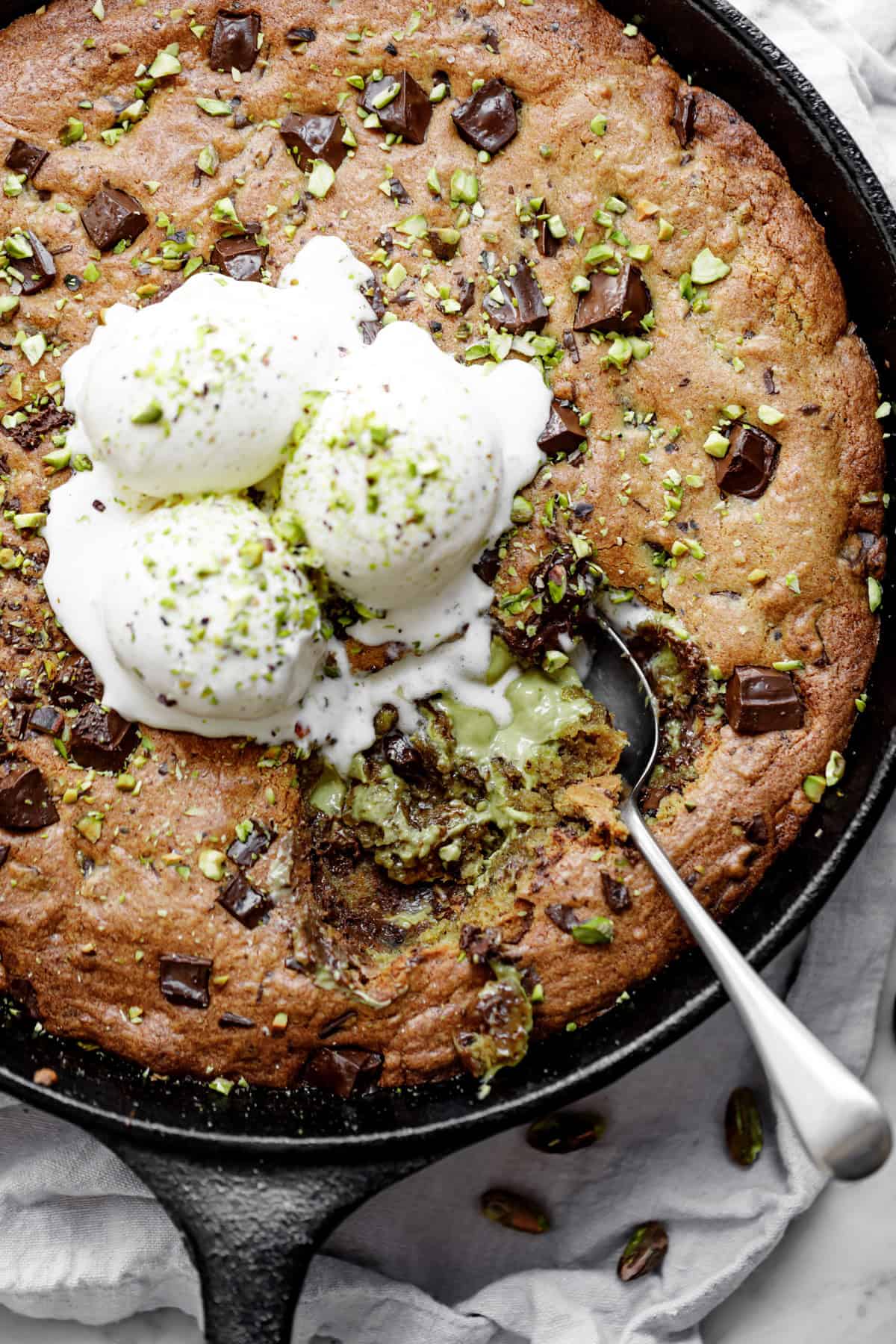 Close-up image of the spoon lifting a portion of pistachio cookie from a skillet, served with vanilla ice cream and topped with chocolate.