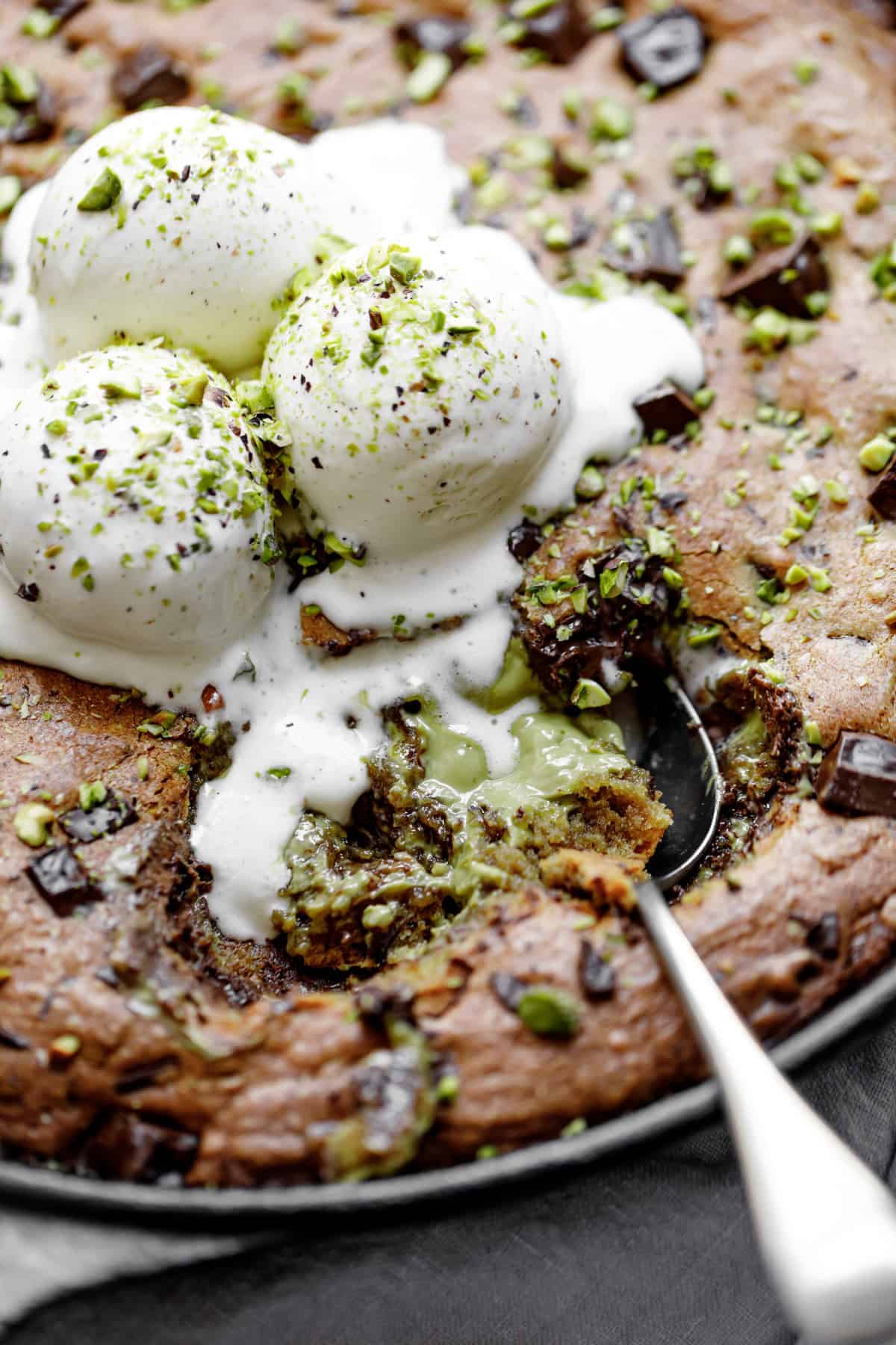 Close-up image of the spoon lifting a portion of pistachio cookie from a skillet, served with vanilla ice cream and topped with chocolate.