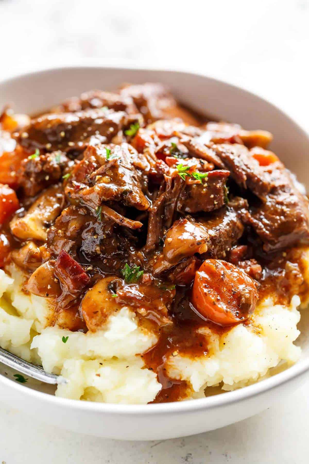 Beef Bourguignon on mashed potatoes with gravy served in a white bowl with a silver fork in a white bowl on a white table