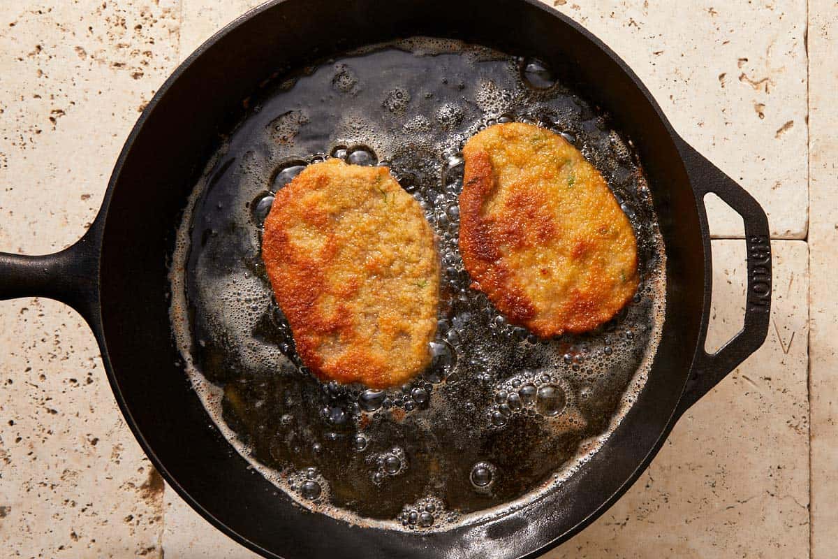 Two beef schnitzels sizzling in hot oil in a black skillet.