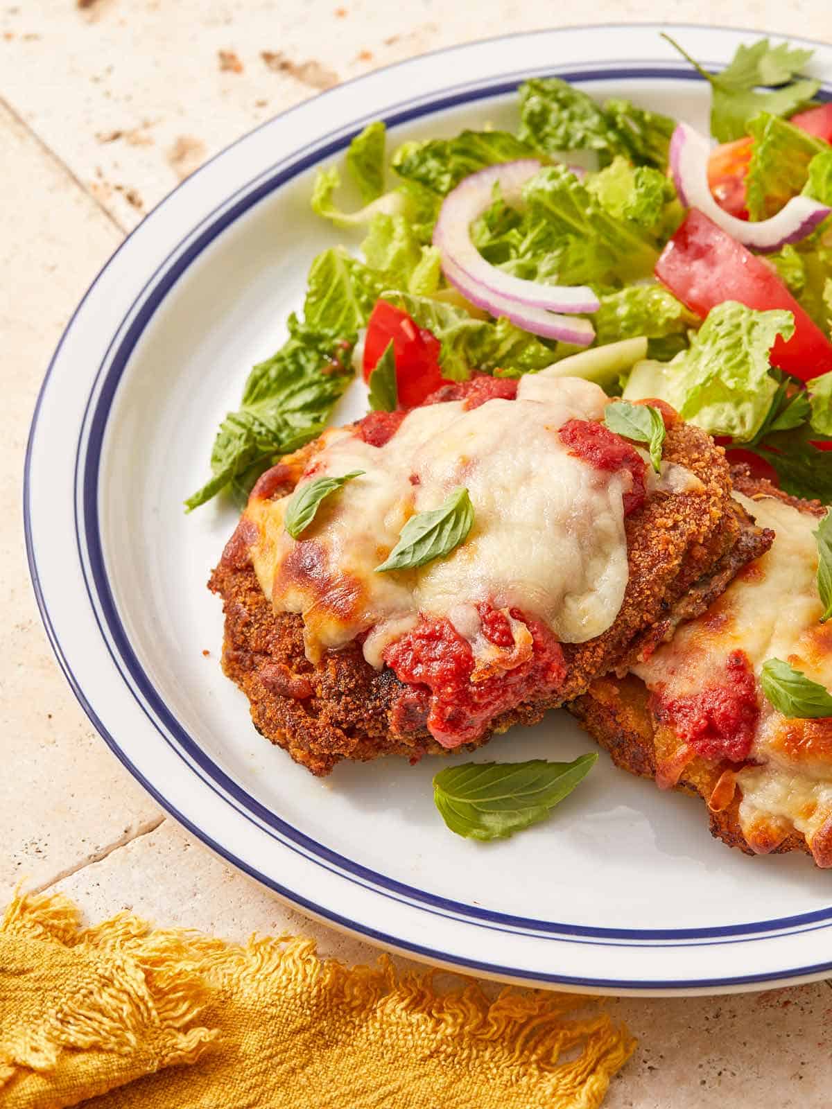 Two crispy beef schnitzels topped with melted mozzarella and tomato sauce, garnished with fresh basil, served alongside a green salad with tomatoes and onion on a white plate.