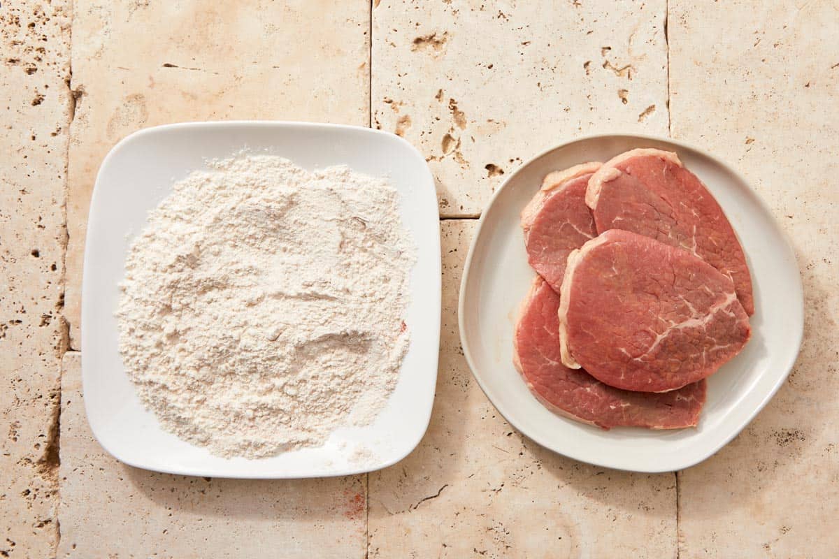 Eye fillet steaks arranged on a plate beside a bowl of seasoned flour on a light stone countertop.