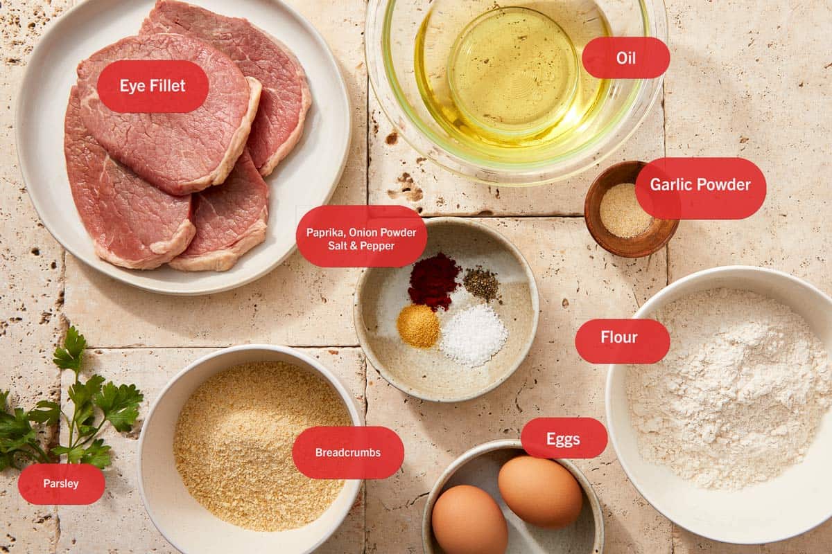 Raw eye fillet steaks, eggs, flour, breadcrumbs, and spices arranged on a wooden board for beef milanesa, labeled 