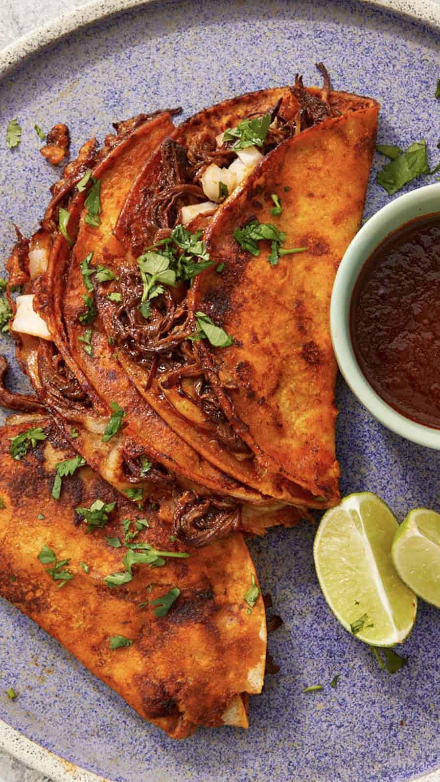 Three Birria Tacos served on a light blue plate with a small bowl of caldo