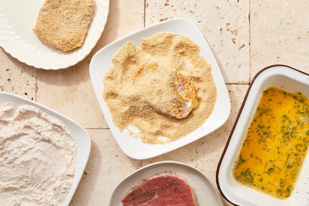 Thin beef slices being breaded step-by-step: flour, egg wash, and breadcrumbs