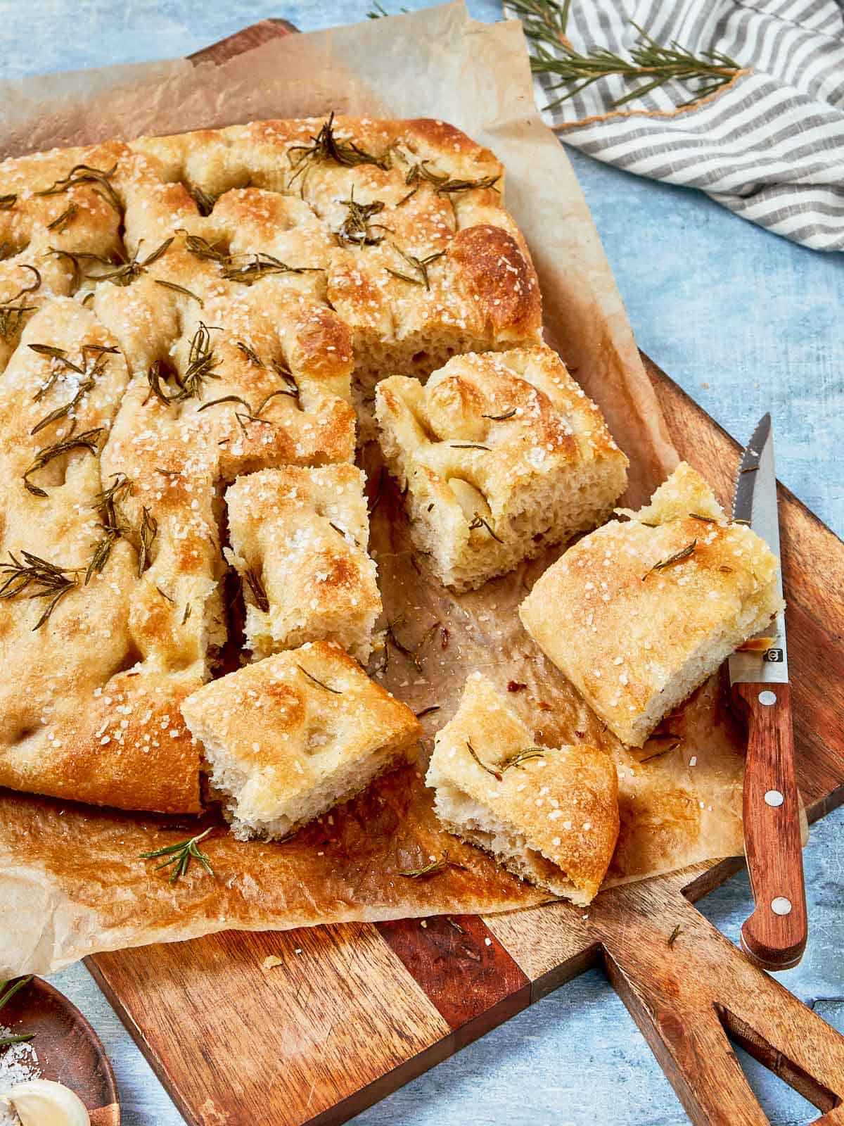 Baked focaccia bread sliced into squares, topped with sea salt and rosemary, served on parchment paper.