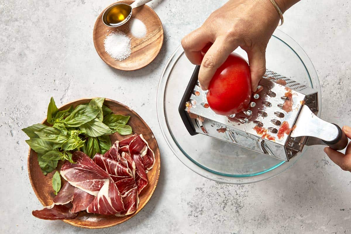 Grating a tomato into a bowl beside basil, Jamón Ibérico, and seasoning ingredients.