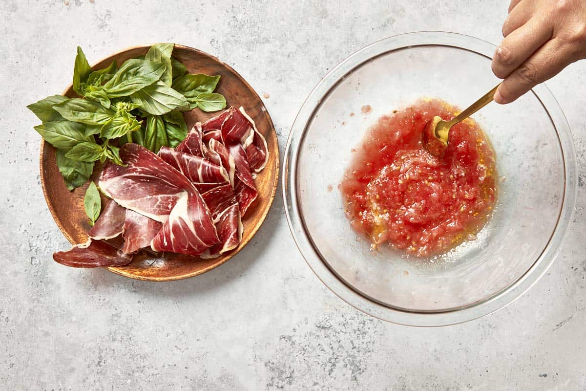 Spanish tomato topping being stirred in a bowl with Jamón Ibérico and fresh basil on the side.