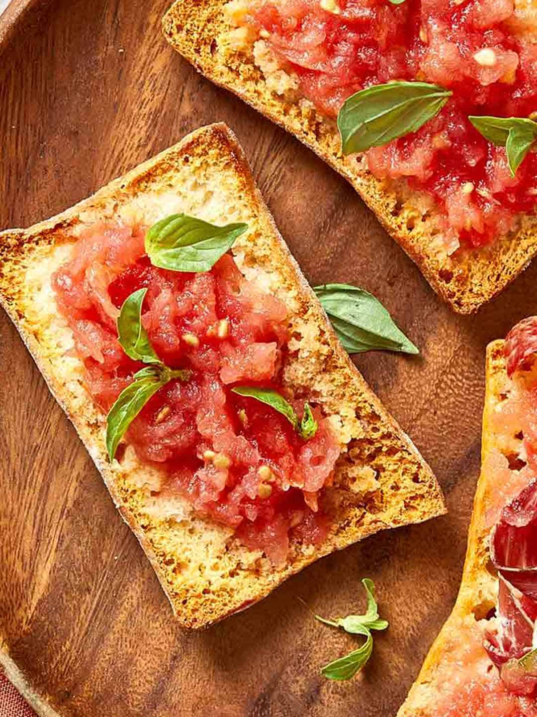 Overhead view of Spanish Pan Coca with tomato, basil, and cured ham on toasted bread, styled on a wooden board.