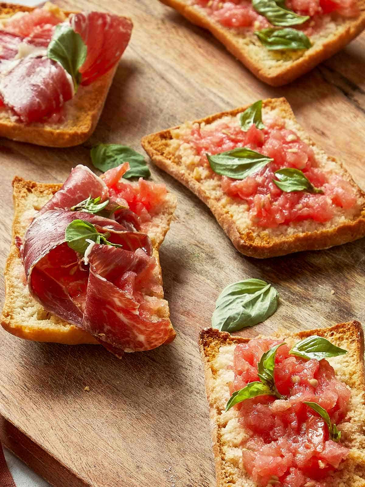 Toasted tomato bread slices topped with grated tomato, fresh basil, and Jamón Ibérico on a rustic wooden board.
