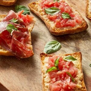 Crispy Pan Coca topped with grated tomato, fresh basil, and slices of Jamón Ibérico on a wooden board
