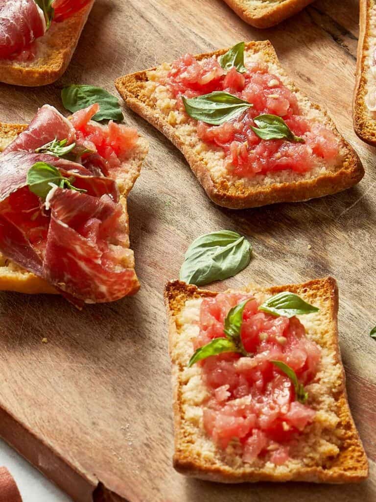 Crispy Pan Coca topped with grated tomato, fresh basil, and slices of Jamón Ibérico on a wooden board
