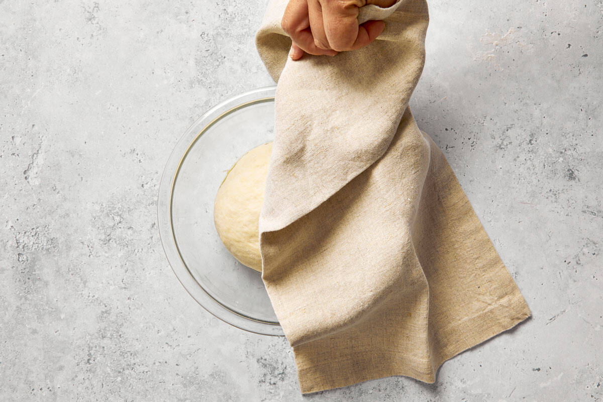 Dough covered with a kitchen towel, rising in an oiled glass bowl.