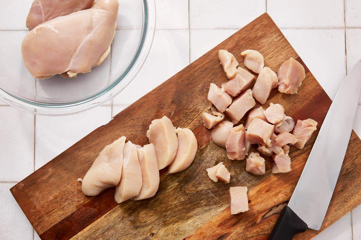 Raw chicken breasts being sliced and diced on a wooden cutting board next to a chef's knife.