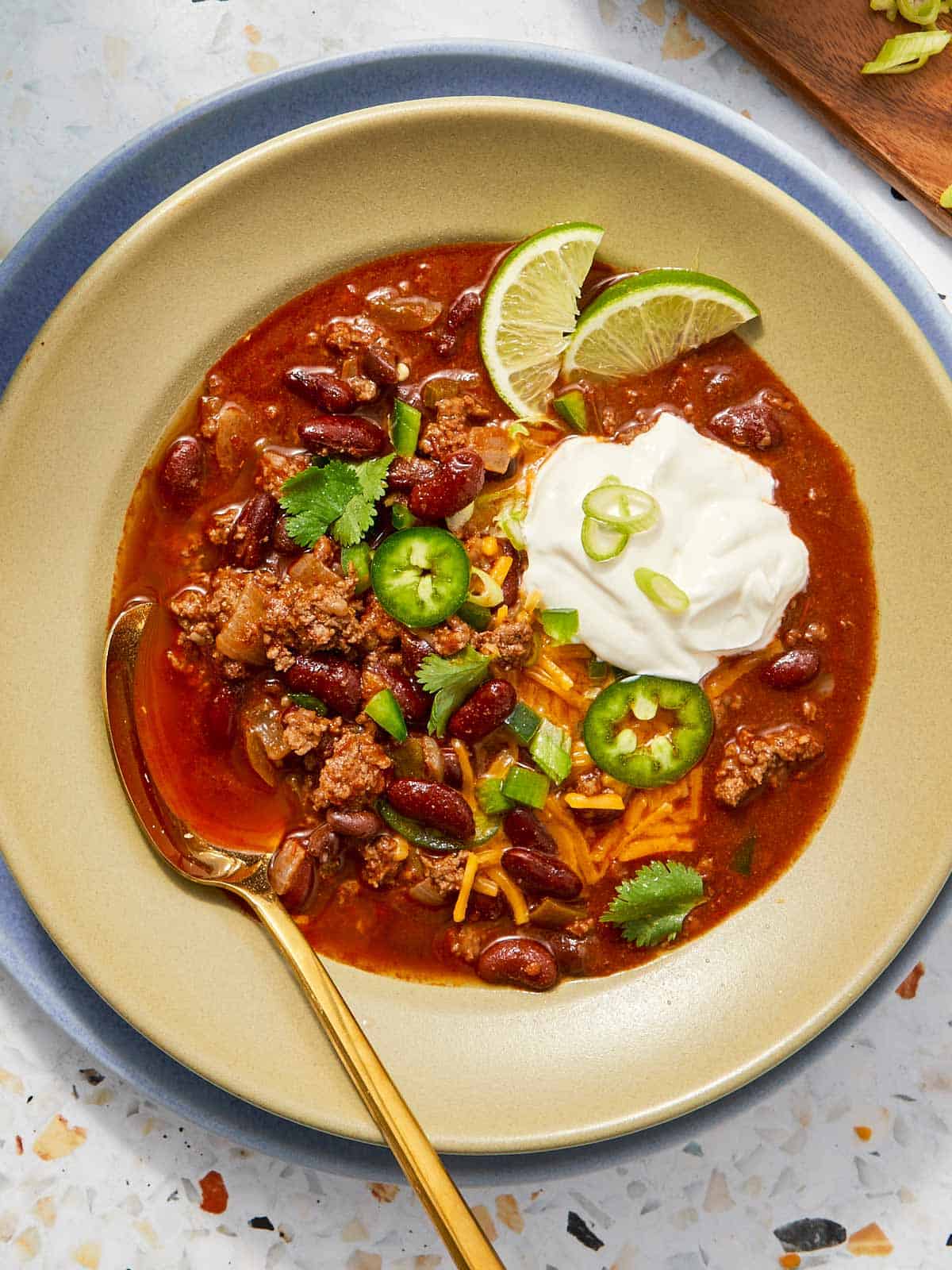 Bowl of beef chili with sour cream, shredded cheese, jalapeños, green onions, and lime wedges.