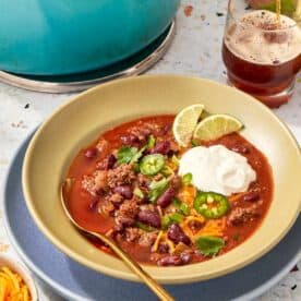 Ladling beef chili into a bowl topped with shredded cheese, cilantro, jalapeños, and lime.