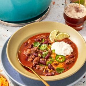 Ladling beef chili into a bowl topped with shredded cheese, cilantro, jalapeños, and lime.